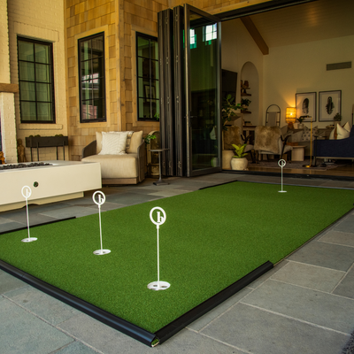 A BirdieBall Outdoor Putting Green—Backyard Tour Grade Synthetic Turf with four numbered flags—is set up on a tiled patio near an open living room featuring modern furniture and decor.