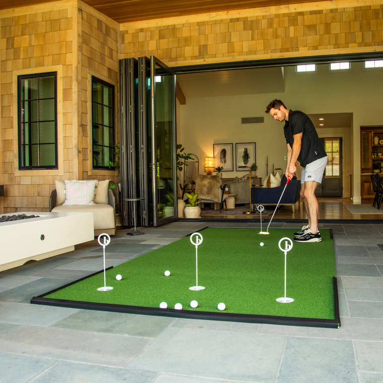 A man practices putting on the BirdieBall Outdoor Putting Green—Backyard Tour Grade Synthetic Turf, with several golf balls scattered on the mat in a spacious living area.