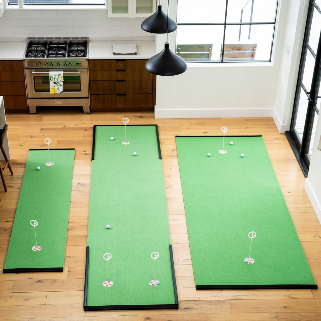 Three BirdieBall Indoor Putting Green - Classic Depth Putting Mats, each with golf balls and flags, are arranged on a wooden floor to create an indoor putting area in a modern kitchen and dining space with large windows.