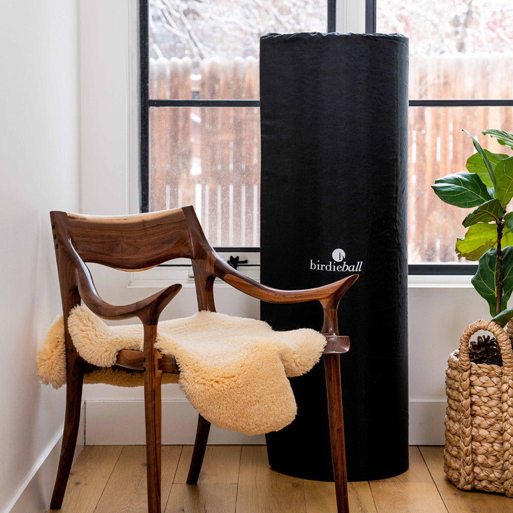 A wooden chair with a cream fleece throw sits beside a tall, black BirdieBall cylinder and The Ultimate Putting Mat Package (4'x10'-4'x18') Indoor RollTech™ Putting Green by BirdieBall in a bright room with a plant and snowy window view.